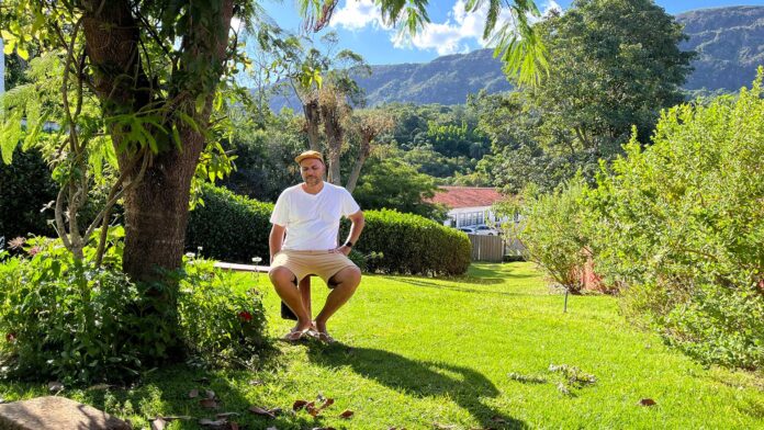 Homem pardo brasileiro sentado num jardim debaixo de uma árvore com uma montanha ao fundo e olhando as formigas no chão.